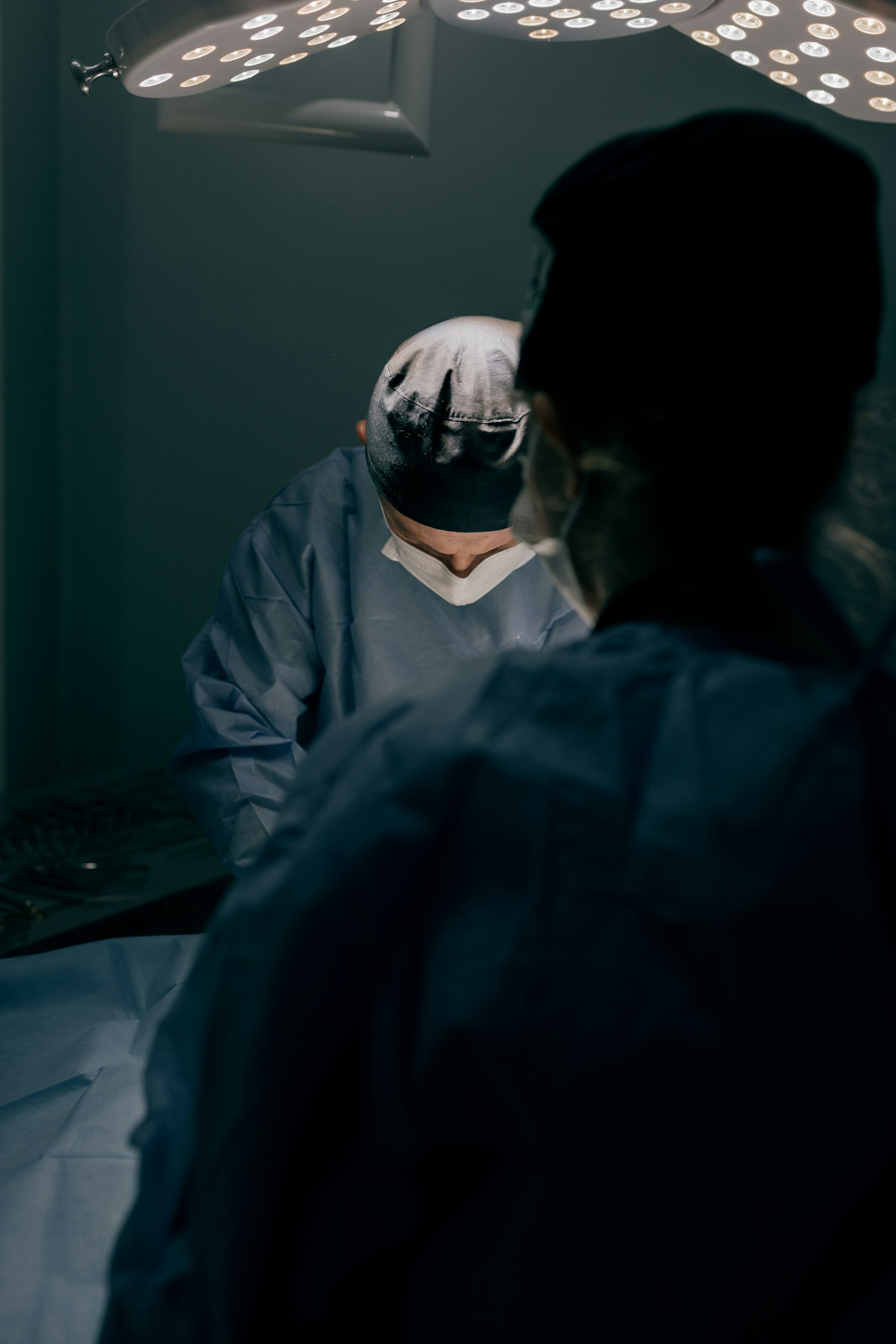 Surgeon working under focused overhead lights, viewed from behind an assistant